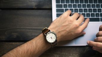 The left hand of a white man wearing a watch on the keyboard of a laptop.  The right hand is slightly in view. 